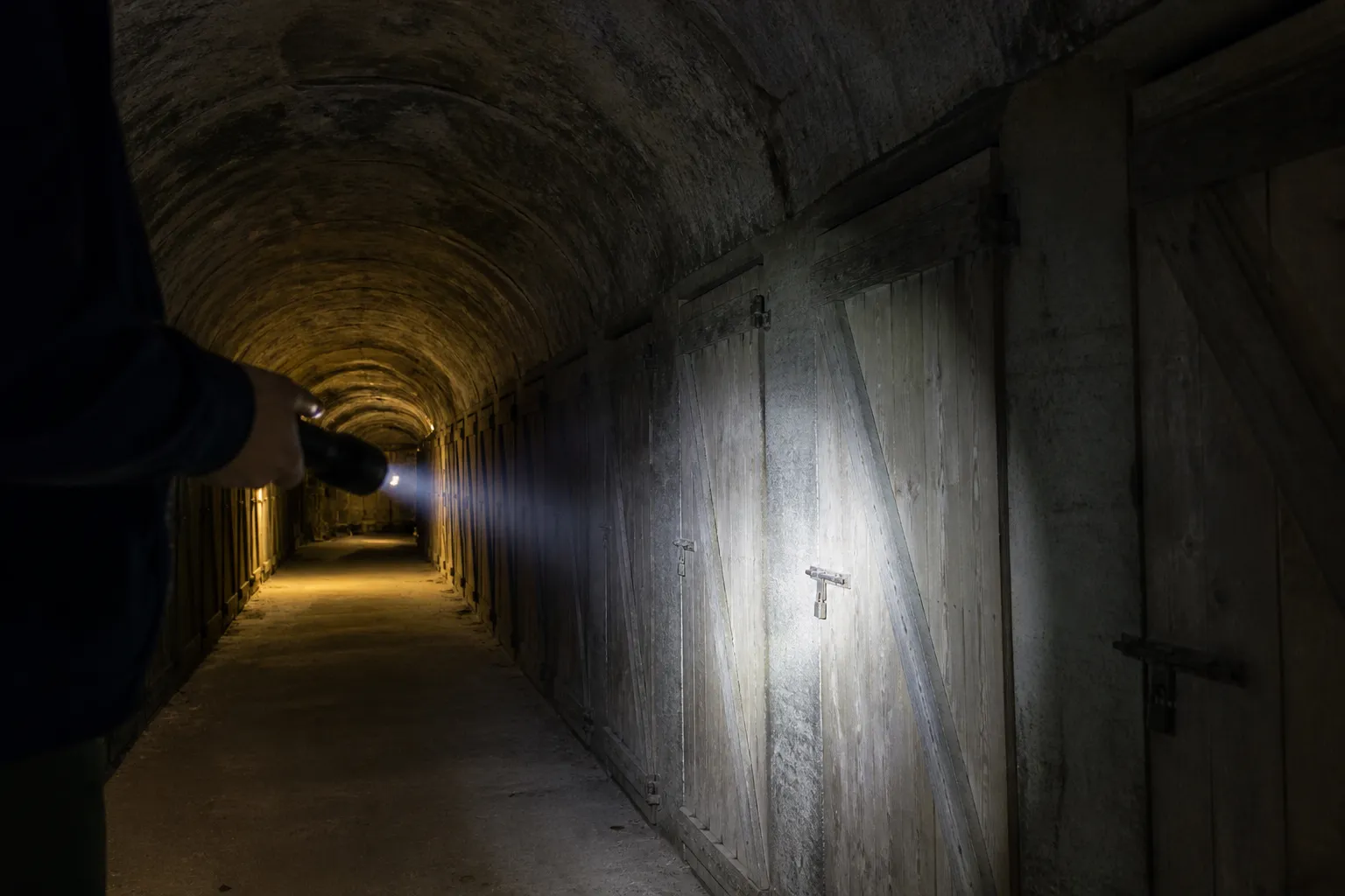 Lampe torche eclairant un couloir de caves voutees avec portes de box en bois